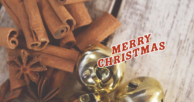 Fototapeta Displaying cinnamon sticks, anise pods, golden bells on wood surface with red Merry Christmas text