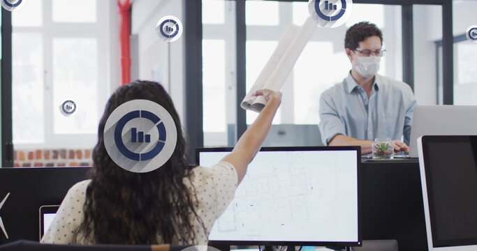 Handing blueprints, designer wearing face mask reaching across monitors in office with infographics - Powered by Adobe