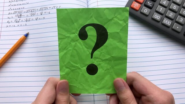A student  holding a green paper note with a question mark on it in his hands over an open notebook with math equations in it and a calculator. Close up.