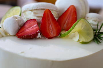 A decorated cake with white frosting is adorned with fresh strawberries, lime slices, meringues, and macarons. This dessert looks festive and appetizing, ideal for special occasions.