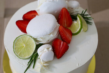 An appetizing cake with white frosting, generously adorned with vibrant strawberry halves, fresh lime slices, crispy meringues, and elegant macarons.