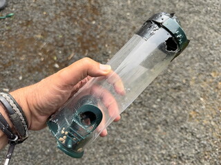 A hand can be seen holding a plastic bird feeder which has had holes chewed in it by a squirrel.