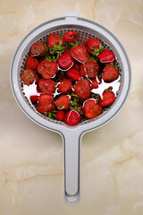 Fresh ripe strawberries in a plastic colander on a marble countertop, top view. Bright red berries with green leaves ready to be washed or used for cooking.