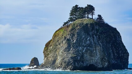 A large rock formation with trees on top surrounded by ocean water under a bright blue sky day light