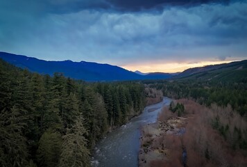 Aerial view of a river flowing through a forest with mountains and cloudy sky in the background