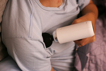 Person using a handheld massage gun on their shoulder while sitting on a couch.