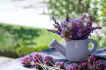 Bouquet of wildflowers in white ceramic watering can. Against backdrop of fresh spring greenery outdoors. For blog about spring delights of country life