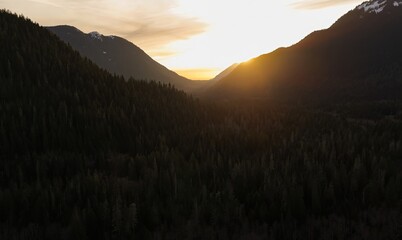 Aerial view of a valley covered in dark trees with mountains and a bright sunset in the distance