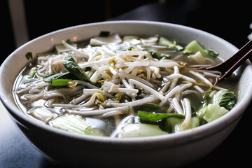 a bowl of Vietnamese pho, seen here with Thai basil and bean sprouts on top of the broth. a spoon is inserted on the side.