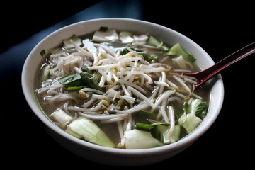 a bowl of Vietnamese pho, Thai basil and bean sprouts on top of the broth. a spoon is inserted on the side.