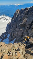 A climber ascending a rocky mountain ridge with snow patches and distant mountain range under a blue sky