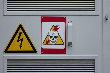 Two Electrical Hazard Warning Signs On A Grey Metal Panel: Yellow Triangle With Lightning Bolt, And...