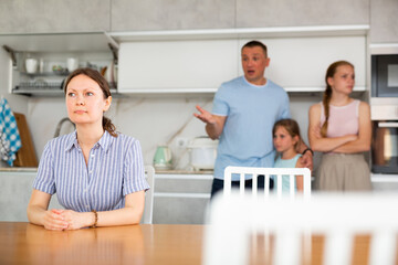 Frustrated, confused woman sits at kitchen table with chin in hands during family conflict. Daughters support father during parents quarrel, husband shouts at his wife