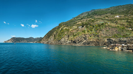 The coast of Cinque Terre with crystal clear water and green terraces on the slopes. A calm bay with a sailboat near the rocky shore creates an atmosphere of Italian harmony.