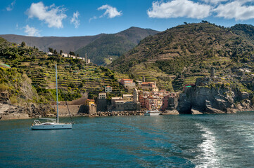 The seaside village of Vernazza, Cinque Terre, shines with colorful buildings, towers fortress nestled between terraced hills, sailboat on the calm blue waters of the Ligurian Sea.