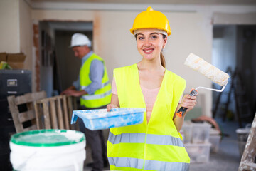 Young woman house painter in protective gear standing on indoor building construction site near...