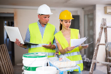 Fototapeta premium Portrait of woman architect and foreman holding blueprints and using laptop while discussing working process in house being renovated