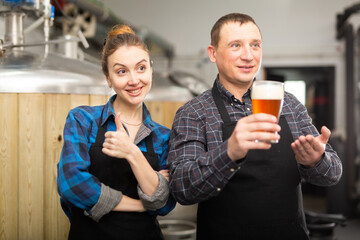 Portrait of positive man and woman brewmasters presenting beer in brewery.