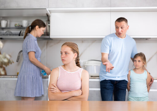 Frowning aggrieved teenage girl sitting at table in home kitchen, listening to reprimand of father standing in background. Concept of family conflicts and misunderstanding of children - Powered by Adobe