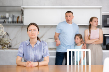 Tense family scene. Upset mom sits in kitchen as husband berates her. Kids side with dad and hug dad