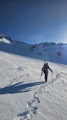 A person hiking up a snowy mountain with skis strapped to their back under a clear blue sky day