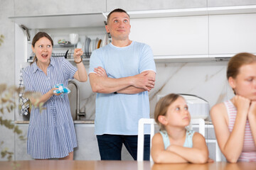 Fototapeta premium Couple sort things out and yell at each other in kitchen. Parents quarrel behind back of their uneven-aged daughters. Children are blurred unrecognizable.