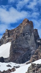 A towering rock formation with snow patches under a bright blue sky dotted with fluffy white clouds