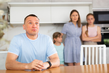 Upset father is sitting at the table, in background is a sad wife with small children. Family quarrel in home kitchen