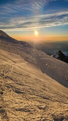 A snowy mountain slope with a bright sun setting on the horizon and a blue sky with clouds above