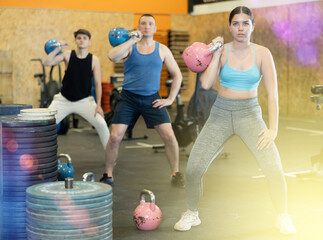 Naklejka premium Concentrated athletic young woman taking part in intense group training in modern gym, performing exercises with kettlebells. Fitness and bodybuilding concept..