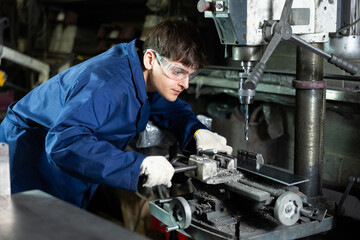 Focused young worker in blue overalls and protective goggles operating industrial drill press in workshop, showcasing concentration and technical skills for holing metal workpiece