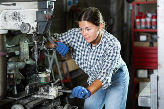 Focused young workwoman in casual plaid shirt and jeans operating industrial drill press in workshop, showcasing concentration and technical skill for holing metal workpiece - Powered by Adobe