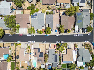 Vertical aerial view of a suburban residential neighborhood with single-family homes, solar panels, driveways, cars, and backyards including a swimming pool and garden 