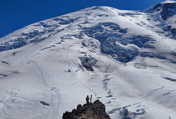 A climber stands on a rocky outcrop with skis against a backdrop of a snow covered mountain and blue sky