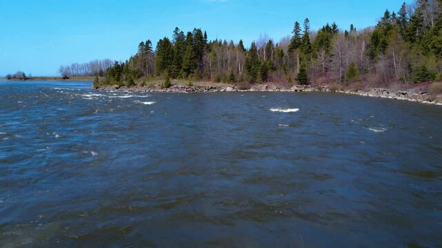 Low-level flight over a river in spring flood to a rocky bank topped with forest under a cloudless blue sky. Mitis River, Quebec, Canada, 2025.