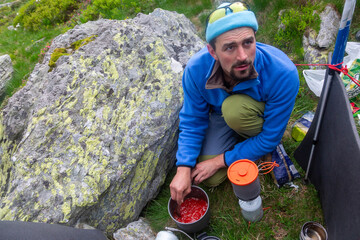 Hiker cooking meal at rocky campsite surrounded by moss-covered boulders. Man in blue jacket preparing food outdoors during wilderness adventure