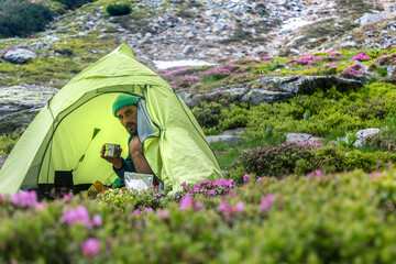 Camper enjoying morning coffee in green tent surrounded by pink wildflowers. Hiker relaxing at scenic mountain campsite with blooming heather.