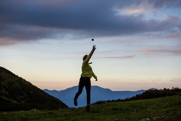 Hiker playing catch on mountain ridge during golden hour, with layered peaks and colorful sky creating perfect backdrop for outdoor activity.