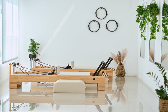 Bright and airy empty pilates studio without people. Beautiful space featuring wooden machine, white design, mirrors and high end fitness equipment with soft sunlight.           