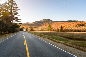 Deserted straight stretch of a road through a valley surrouned by forested mountains at the peak of autumn colours at sunset