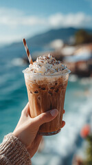 Hand holding refreshing iced coffee with whipped cream and chocolate powder topping, enjoying a sunny day by the sea