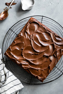 A square chocolate cake with rich frosting on a cooling rack, surrounded by baking tools and a striped cloth.