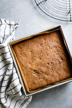 A freshly baked square cake in a parchment-lined baking pan on a light gray surface, with a striped kitchen towel and a cooling rack nearby.