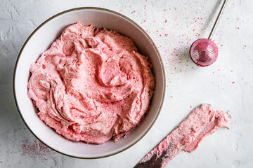 A bowl of pink whipped cream with a spatula and sieve on a textured white surface.