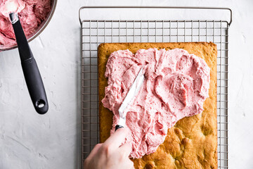 A person spreads pink frosting on a rectangular cake using a spatula on a cooling rack.
