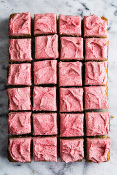 An overhead view of a batch of square cake slices with pink frosting arranged on a marble surface.