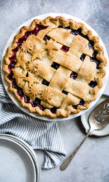 A freshly baked berry pie with a lattice crust and decorative star shapes on top, placed on a striped cloth with a pie server beside it.
