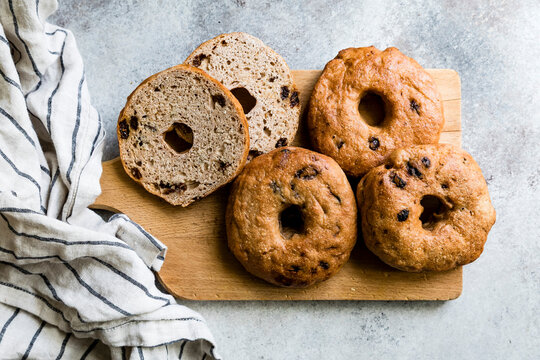 A wooden board with four bagels, three whole and one sliced, next to a striped kitchen towel.