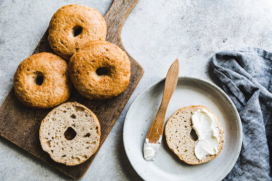 An assortment of bagels on a wooden board with one sliced and spread with cream cheese on a plate.