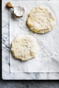 Two portions of dough on a floured parchment paper with a wooden scoop of flour on a marble surface.